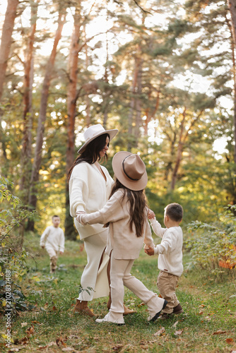 A mother and her children hold hands and play during a golden autumn walk through a sunlit forest, wearing cozy neutral sweaters and wide-brim hats, conveying warmth, connection and family 