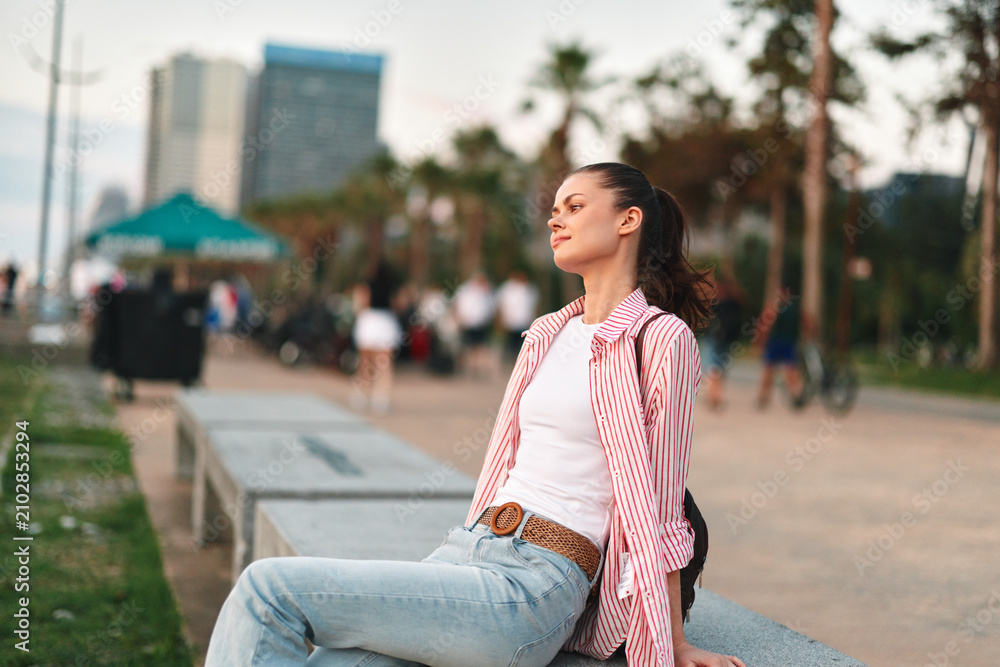Fototapeta premium Young woman with long dark hair sits on a concrete bench wearing a white tee and striped shirt, relaxed in a sunny park with a distant city skyline.