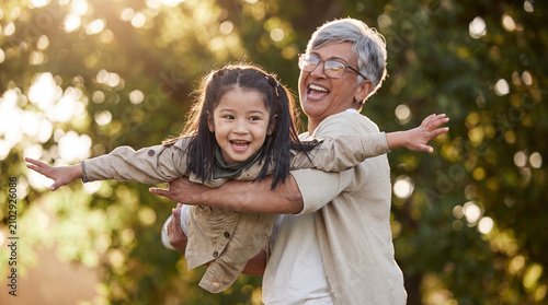 A happy grandmother lifting her granddaughter in the air like an airplane during a sunny afternoon at the park, showcasing love and multi-generational bonding.