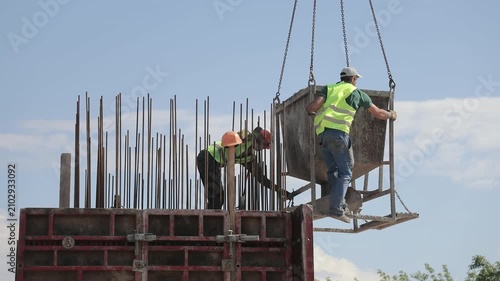 Construction workers guide a suspended concrete hopper above formwork, Crane chains and exposed reinforcing bars show active building operations