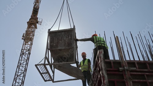 Construction workers guide a suspended concrete hopper beside tall formwork, Tower crane and exposed reinforcing bars frame an active building site