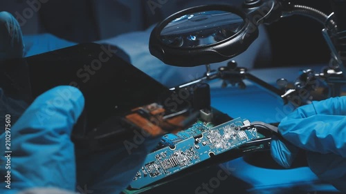 Technician inspecting a circuit board with a magnifying glass. Close up of hands in blue gloves working on microelectronics in a laboratory.