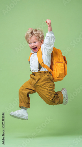A cheerful young boy in autumn-colored clothing and a bright yellow backpack leaps through the air with his fist raised in excitement.