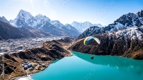 Paraglider soaring over a stunning turquoise lake with snow-capped mountains in the background. A village nestled near the shore adds to the breathtaking landscape