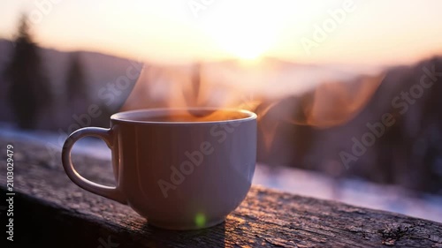 A steaming cup of coffee sits peacefully on a rustic wooden railing, as golden sunlight gently rises above majestic mountains, illuminating the tranquil morning scene with warmth.
