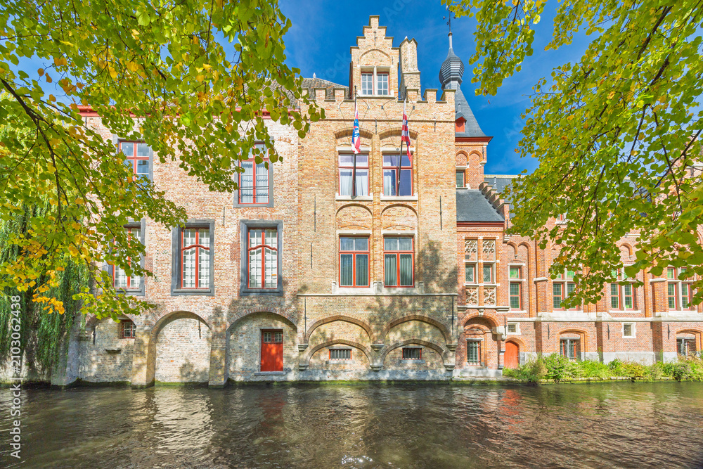 Fototapeta premium Bruges town in Belgium, Europe. Historic European Canal View with Ancient Architecture and Lush Green Foliage on a Bright Sunny Day, Reflecting Tranquil Waters Below