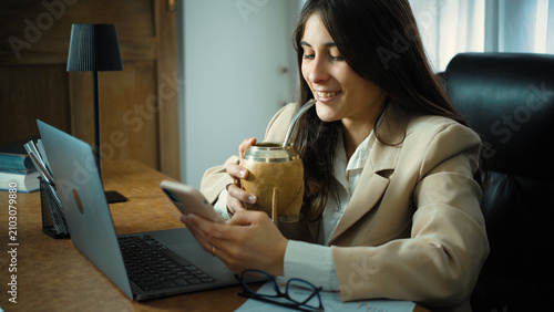 Konstfotografi Young Hispanic businesswoman taking a relaxing break in her office while drinking yerba mate and using her smartphone, enjoying a peaceful moment during her workday in a modern workplace