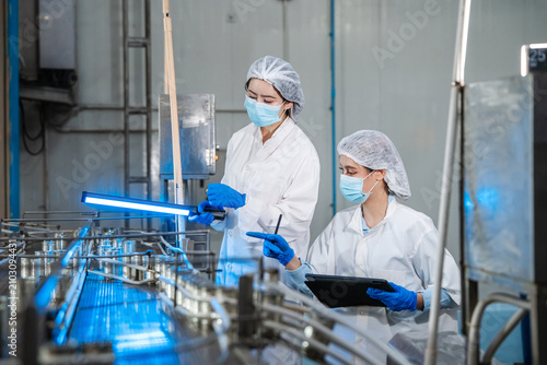 Two women in white lab coats inspect machinery. Blue LED lights illuminate metal conveyor systems. One holds a tablet, the other a pen. They wear hairnets and gloves. Factory setting with pipes
