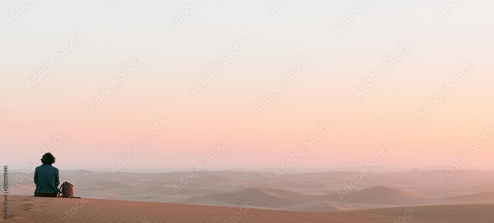 Fototapeta premium Solo Traveler Resting on Desert Rock Brushing Hiking Boots After Trek, Sculpted Sand Dunes at Sunset with Expansive Copy Space and Authentic Documentary Travel Photography