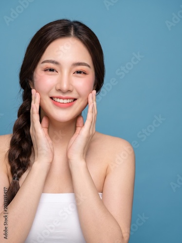 young woman with long dark braided hairstyle, natural makeup, wearing a white strapless tube top, touching both cheeks, with a happy smile, isolated on a blue seamless studio background