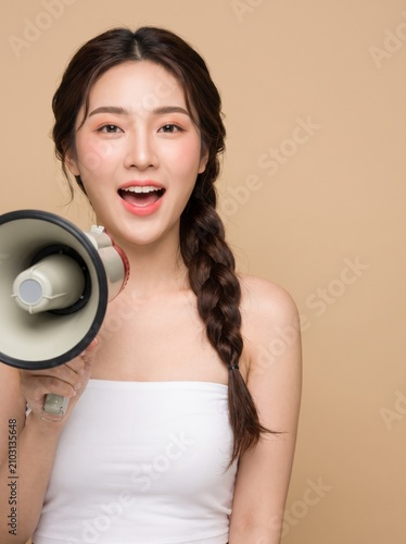 young woman with long dark braided hairstyle, natural makeup, wearing a white strapless tube top, holding a megaphone, with an excited expression, isolated on a beige seamless studio background