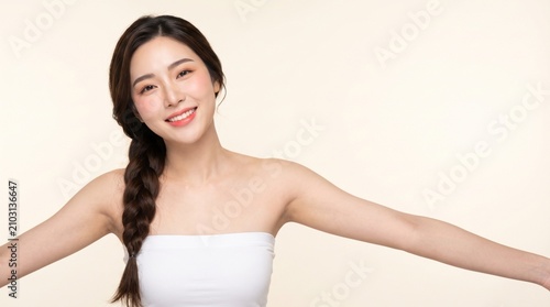 young woman with long dark braided hairstyle, natural makeup, wearing a white strapless tube top, opening both arms, with a warm smile, isolated on a cream seamless studio background
