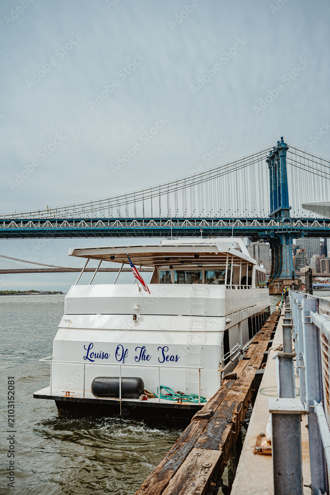Fototapeta premium New York, USA, 2 May 2026: White cruise ship docked on East River near Manhattan Bridge. Back side of tour boat at the pier with Manhattan Bridge and Brooklyn Bridge visible in distance.