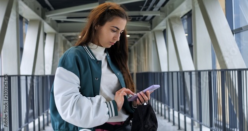 Young woman offering friendly glance in pedestrian corridor, slight smile while holding phone and bag, confident posture with relaxed shoulders, modern geometric background and soft daylight creating