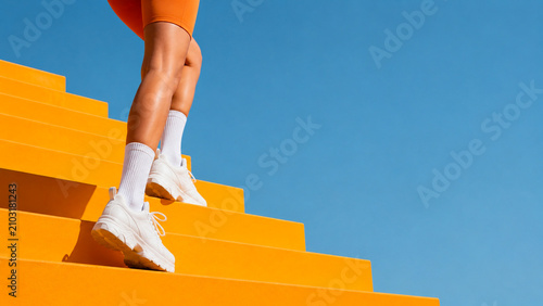 Athletic woman walking up bright orange stairs against a clear blue sky.