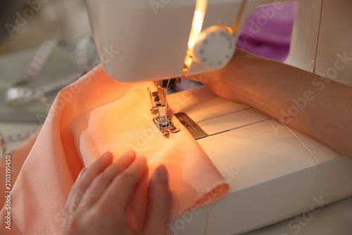 Woman sewing fabric with professional machine at white table in workshop, closeup