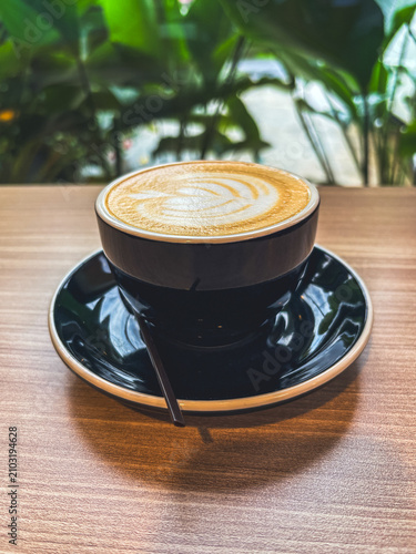 Cozy cup of coffee sits on wooden table. Smooth latte art glows softly above dark ceramic. Green leaves frame background with natural warmth. Serene moment captured in quiet cafe setting