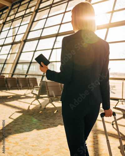 A professional blonde businesswoman walking through a bright modern airport terminal with her suitcase ready for a business trip abroad