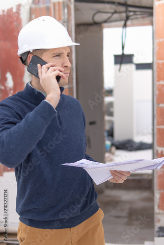 Construction engineer wearing hard hat calling on phone, holding blueprint plans, managing project communication and building development at construction site