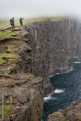 Two hikers standing on the edge of dramatic Trælanípa sea cliffs with one pointing towards the ocean in fog, Faroe Islands