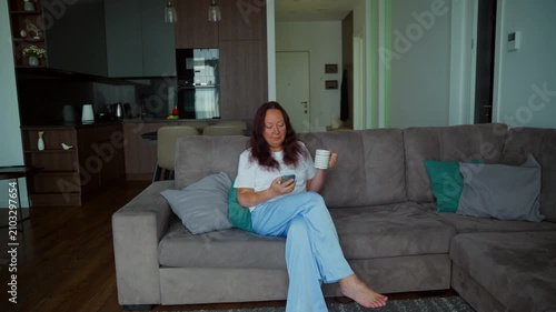 Woman sitting on gray sectional sofa holding coffee cup, scrolling smartphone in contemporary open plan apartment with kitchen visible.