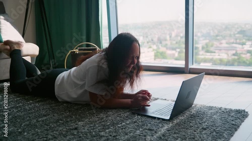 Woman reclines on carpet floor with laptop in modern high rise apartment overlooking sprawling cityscape through floor to ceiling windows, relaxed work posture.