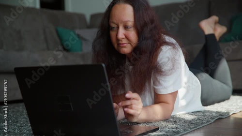 Woman lying on floor indoors, focused on laptop screen while resting near gray couch in comfortable home setting.