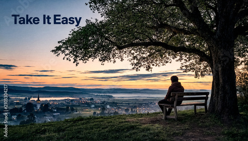 Silhouette of a person sitting alone on a wooden bench under a large tree overlooking a valley town at sunset and text Take It Easy.