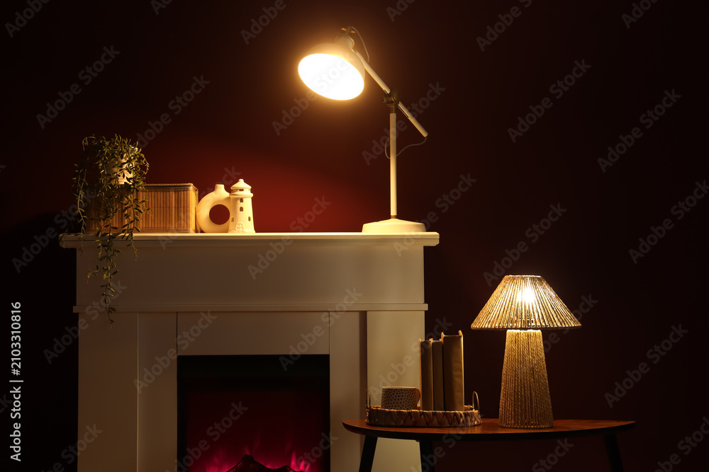 Obraz premium Interior of stylish room with fireplace, coffee table and glowing lamp near red wall at night