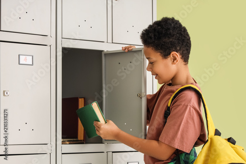 Teenage African-American schoolboy taking book from locker on green background