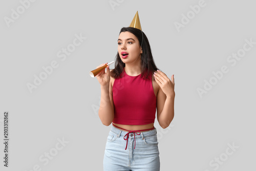 Beautiful young woman in party hat with whistle on grey background