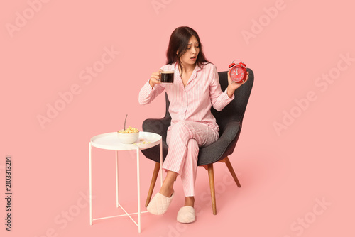 Young Asian woman in pajamas with alarm clock, cup of coffee and cornflakes s...
