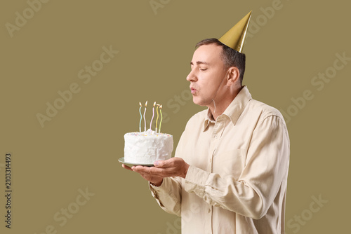 Mature man with birthday cake making wish on green background