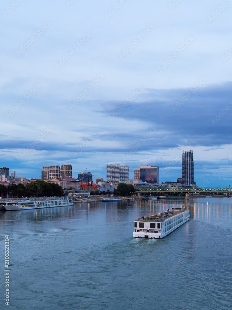 Obraz premium Cityscape with boat on river at dusk. A boat moves along the river as buildings rise above the water in a city at dusk. The sky is filled with clouds.