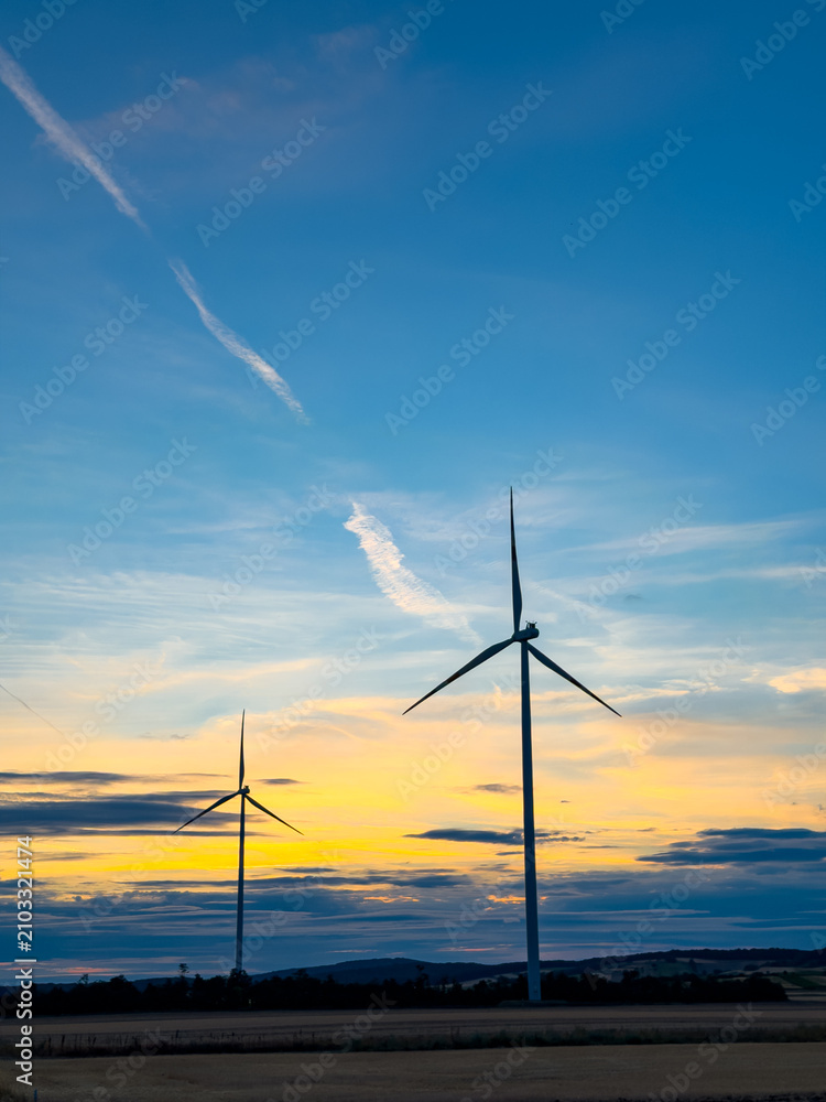 Obraz premium Turbines at sunset. Two wind turbines stand tall against a sunset sky in a rural area. The colorful clouds create a scenic view.