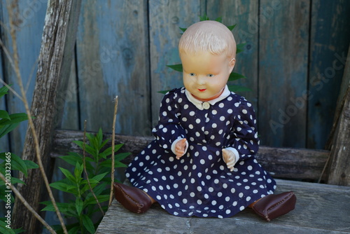 A vintage plastic baby doll wearing a blue polka dot dress sits on a weathered wooden structure against a distressed blue background