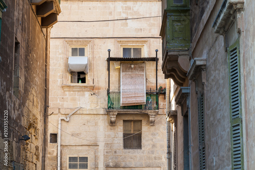 Weathered Valletta buildings with green wooden gallarija balconies and an air conditioner unit line a narrow shaded alley