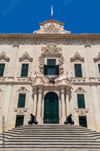 The symmetrical baroque facade of Auberge de Castille rises above curved steps flanked by cannons, with a flag flying atop in Valletta, Malta