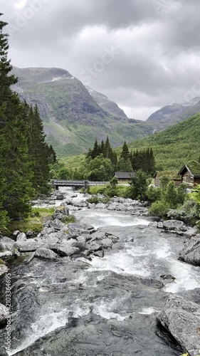 Traditional Norwegian houses with grass-covered roofs nestled beside a mountain river flowing from the surrounding peaks. A picturesque bridge crosses the crystal-clear stream, while forests and majes