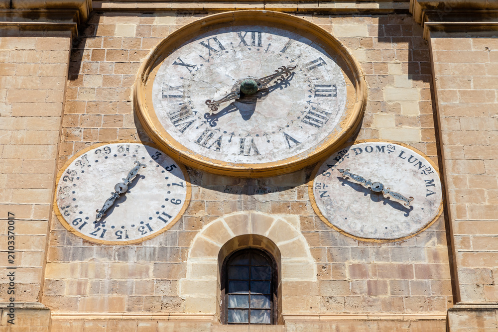 Fototapeta premium An ornate church clock face with Roman numerals, flanked by smaller date and day dials, adorns a stone tower in Valletta, Malta