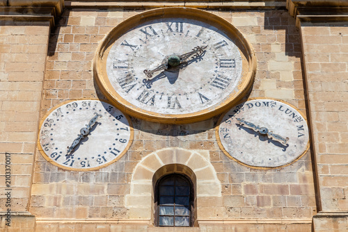 An ornate church clock face with Roman numerals, flanked by smaller date and day dials, adorns a stone tower in Valletta, Malta