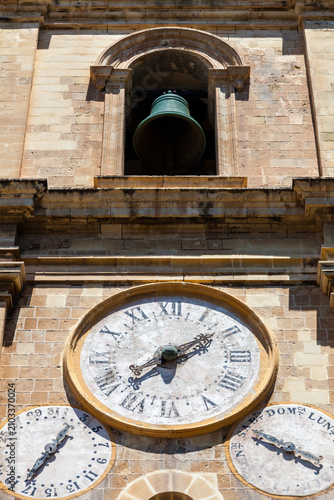 An ornate church clock face with Roman numerals, flanked by smaller date and day dials, adorns a stone tower in Valletta, Malta
