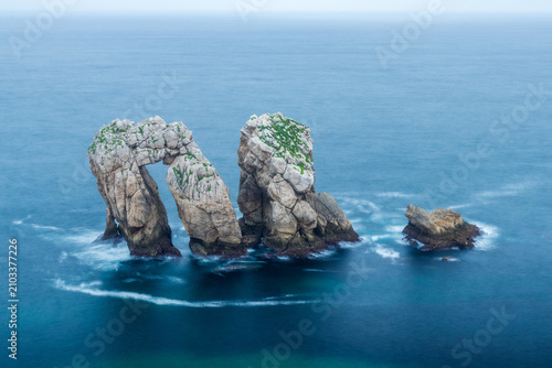 Iconic Urro del Manzano rock formation in the Costa Quebrada Geopark, Cantabria, Spain, on a tranquil summer day with calm seas and cloudy skies.