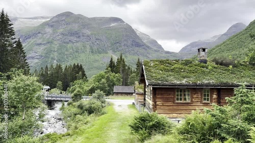Scenic mountain landscape along the famous Trollstigen road in Norway featuring traditional grass-roof houses, a crystal-clear river flowing through the valley, lush forests, and majestic mountains. B