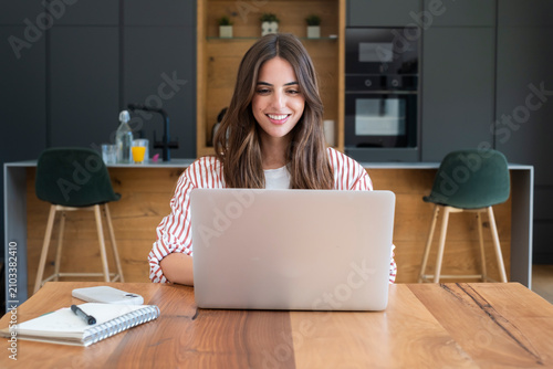 Young woman smiling while working on a laptop at a wooden kitchen table, remote work from home concept, casual home office, notebook and coffee nearby, productivity and focus