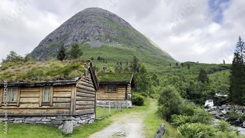 Scenic mountain landscape along the famous Trollstigen road in Norway featuring traditional grass-roof houses, a crystal-clear river flowing through the valley, lush forests, and majestic mountains.