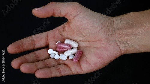 Hand holding various pills and capsules against a dark background. A person's open palm displays a collection of white and pink pharmaceutical capsules and tablets symbolizing medication and treatment