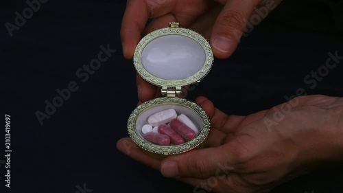 Hands holding an ornate pillbox filled with various capsules and tablets, a collection of different sized and colored pharmaceutical pills