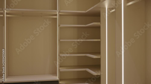Empty walk in closet with built in shelves and hanging rods. LED lights glow along edges. White walls and black frames create clean, modern space with organized layout.