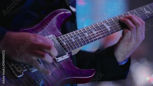 Close up of person playing purple electric guitar with bird inlays. Hands press frets and strum strings. Blurred background suggests stage or music setting with lights.